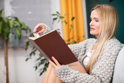Blonde woman during relaxation in living room. During leisure time at home, a woman with shapely legs is browsing through a book while sitting on the sofa.