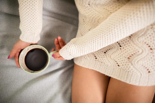 Close-up frame. A woman with slim legs sits on a bed in a living room and next to her holds a porcelain cup with hot coffee.