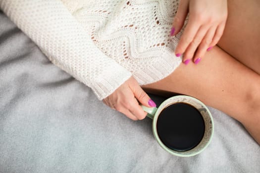 Woman holds coffee cup close to her. Slender female legs with pale complexion. Relaxing in the comfort of home.