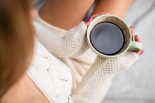 View from above as a woman in a cream sweater holds a delicately porcelain cup with hot coffee. Love coffee and relaxing in the comfort of your home.