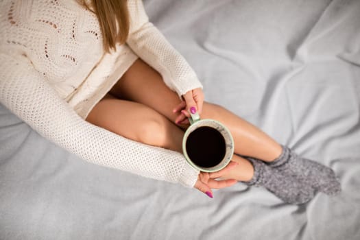 Woman with attractive legs sits on sofa and in her hands holds porcelain cup with morning coffee. A moment of relaxation and rest in the comfort of home.