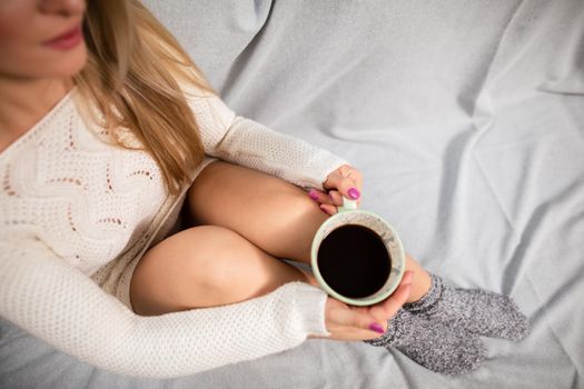 A woman with shapely legs sits on a sofa and holds a porcelain cup with hot coffee in her hands. A moment of relaxation and rest in the comfort of home.