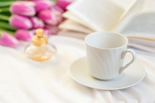 A spring still life with a white mug and a saucer, a golden bottle of perfume, an open book and a bouquet of beautiful pink tulips lies on a table on a white tablecloth. close-up. copy space