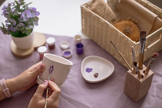 Womens hands paint a white flower ceramic pot with a brush, draw a sprig of lavender. On the table are purple craft paper, brushes, paint jars, and a wicker box. Close-up. View from above