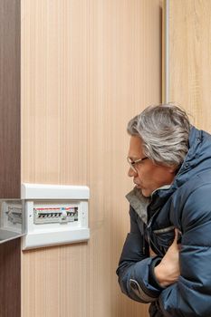 An adult man in a warm jacket looks at the electrical panel. Energy saving concept. Expensive electricity.