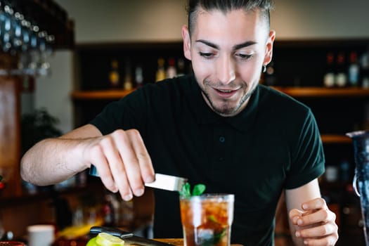 Young and modern waiter, with long dark hair, dressed in black polo shirt, placing mint leaves to decorate a Mojito. Waiter preparing a cocktail. Cocktail glass with ice cubes. Mojito. Bar full of cocktail ingredients. Dark background and dramatic lighting. Horizontal view.