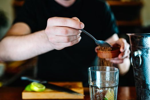 Young and modern waiter, with long dark hair, dressed in black polo shirt, adding brown sugar in a large crystal glass to prepare a cocktail. Waiter preparing a cocktail. Cocktail glass with ice cubes. Mojito. Bar full of cocktail ingredients. Dark background and dramatic lighting. Horizonta