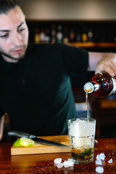 Young and modern waiter, with long dark hair, dressed in black polo shirt, adding liquor in a crystal glass full of ice to prepare a Mojito. Waiter preparing a cocktail. Cocktail glass with ice cubes. Mojito. Bar full of cocktail ingredients. Dark background and dramatic lighting. Vertical view