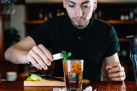 Young and modern waiter, with long dark hair, dressed in black polo shirt, placing mint leaves to decorate a Mojito. Waiter preparing a cocktail. Cocktail glass with ice cubes. Mojito. Bar full of cocktail ingredients. Dark background and dramatic lighting. Horizontal view.