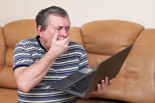 A man with a surprised face holds a laptop in his hands while sitting on the couch. Emotions from the news