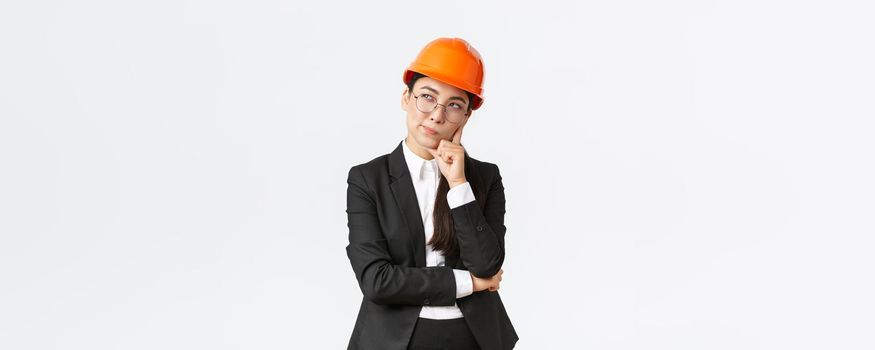 Thoughtful creative female asian chief architect, construction engineer thinking, wearing safety helmet and suit, pondering best choice for building, standing white background.