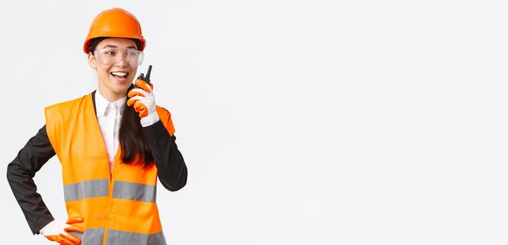 Confident pleased asian female engineer in safety helmet and uniform talking with chief architect using walkie-talkie. Satisfied construction technician contact team using radio phone.