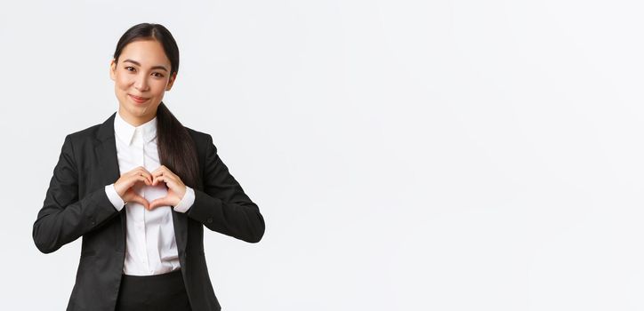 Close-up of pleasant asian businesswoman, saleswoman in black suit care for her clients, showing heart sign and smiling with admiration, standing white background caring and tender.