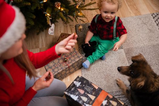 Under mom's supervision, son feeds treats to dog. Mom sits contentedly on the floor next to the Christmas tree.