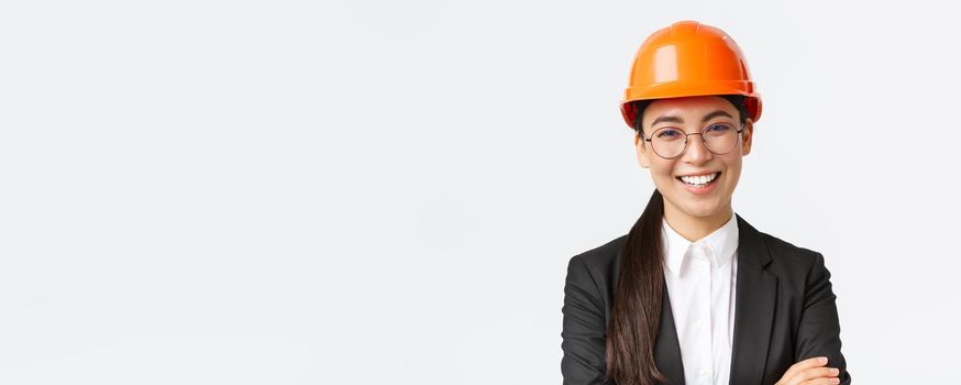 Smiling leader of the team, asian female chief engineer in safety helmet and suit cross arms confident, smiling happy at camera, introduce enterprise, standing white background.