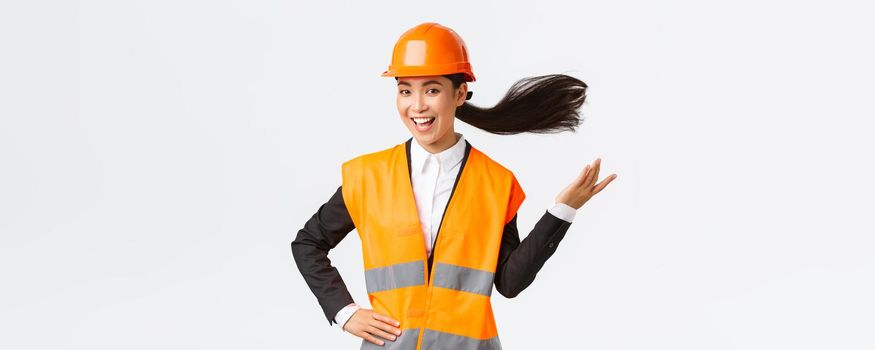 Successful and confident asian female chief engineer, architect in safety helmet looking upbeat, winning and triumphing as finish great project, feeling superior, standing white background.