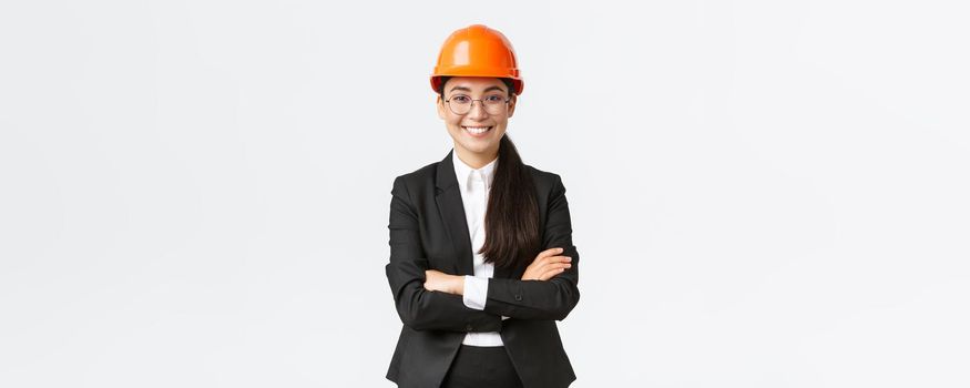Professional smiling asian businesswoman in safety helmet and suit cross arms and looking confident, selling new buildings. Construction manager, engineer showing around, greeting investors.