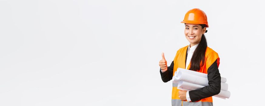 Confident successful female asian construction manager, architect in safety helmet and jacket, showing thumb-up and carry blueprints of building project, guarantee quality, white background.