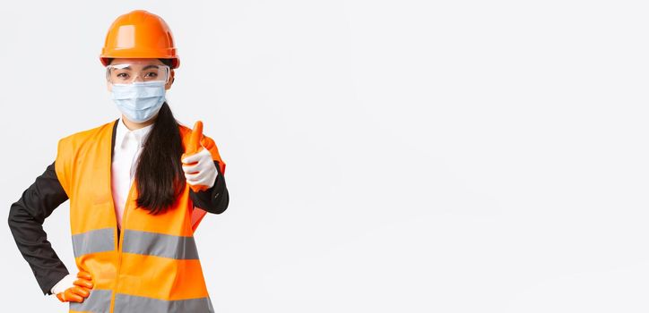Covid-19 safety protocol at enterpise, construction and preventing virus concept. Confident asian female engineer, architect in face mask and helmet showing thumb-up, guarantee quality of building.