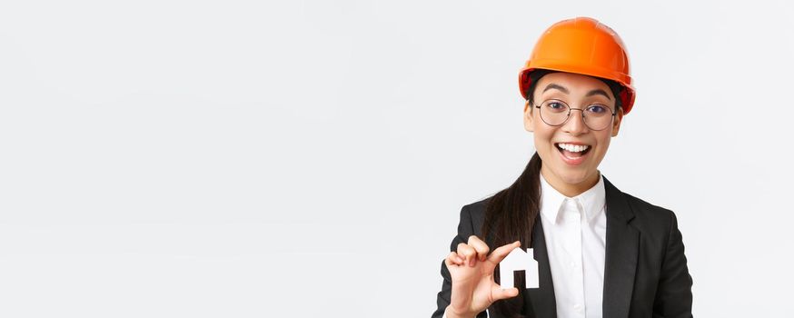 Close-up of professional broker in safety helmet and business suit selling homes. Cheerful architect showing minuature of house and smiling, working over new design or construction, white background.