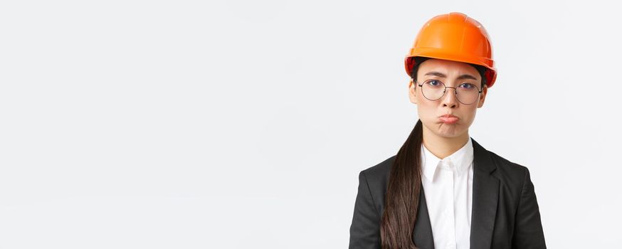 Close-up of upset and gloomy asian female construction manager facing failure, bad results on factory, engineer standing in safety helmet and business suit pouting sad, standing white background.