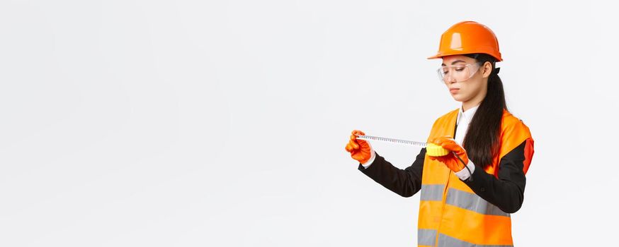 Serious asian female construction engineer, technician inspect layout, measuring something, looking at tape measure with focused face, standing over white background in safety uniform.