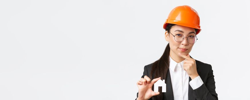 Thoughtful creative female asian architect in helmet and business suit creating new ideas of design, showing home maket and thinking, making cunning face, pondering plan of construction.