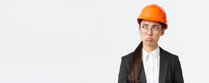 Close-up of gloomy upset asian female engineer in safety helmet and business suit complaining, looking disappointed, whining with sulking face, unwilling work on weekends, looking left bothered.