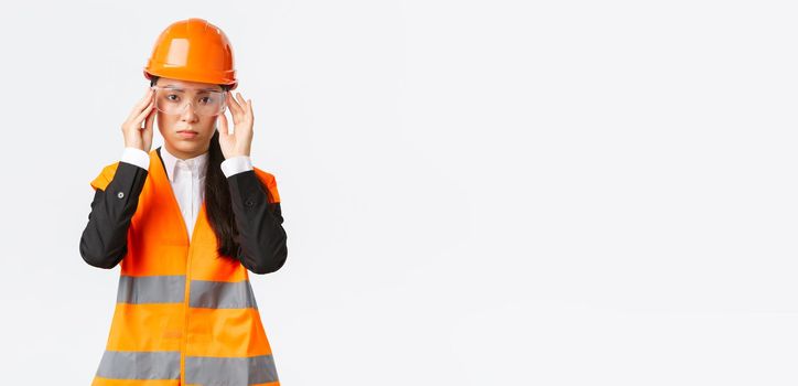 Serious-looking young asian female construction manager follow safety protocol, wearing protective glasses and helmet before entering enterprise, standing white background.