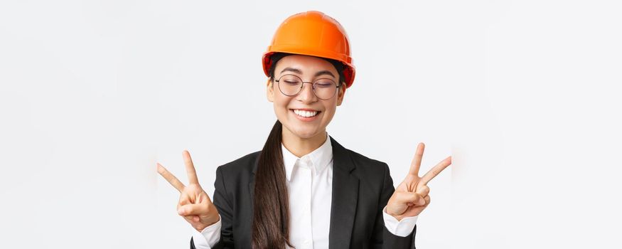 Close-up of cheerful successful female asian engineer, construction architect in safety helmet and business suit showing peace signs and smiling kawaii, standing white background.