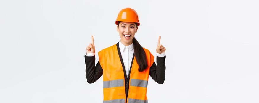 Building, construction and industrial concept. Happy smiling female asian engineer in safety helmet and reflective clothing, introduce new object, estate for sale. Architect pointing fingers up.