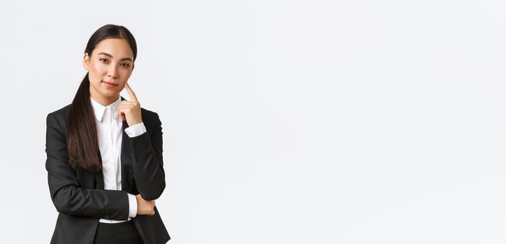 Creative smart asian female manager, saleswoman in suit smiling cunning and looking at camera, have plan, thinking, got excellent idea, standing pleased over white background.