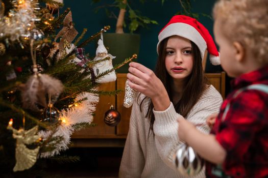 A sister with her younger brother decorates a Christmas tree. A two-year-old child with curly blonde hair.