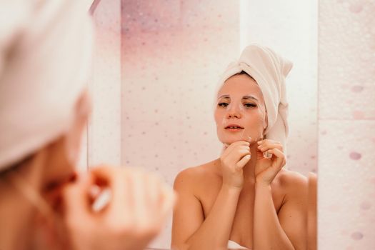 Young beautiful woman using a moisturizing facial mask after taking a bath. Pretty attractive girl in a towel on her head stands in front of a mirror in a home bathroom. Daily hygiene and skin care.