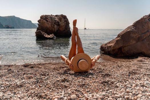 A beautiful middle-aged woman lies on the beach with her feet to the sky, covering her body with a wide-brimmed straw hat. She is sunbathing on the ocean. Vacation, travel, vacation concept