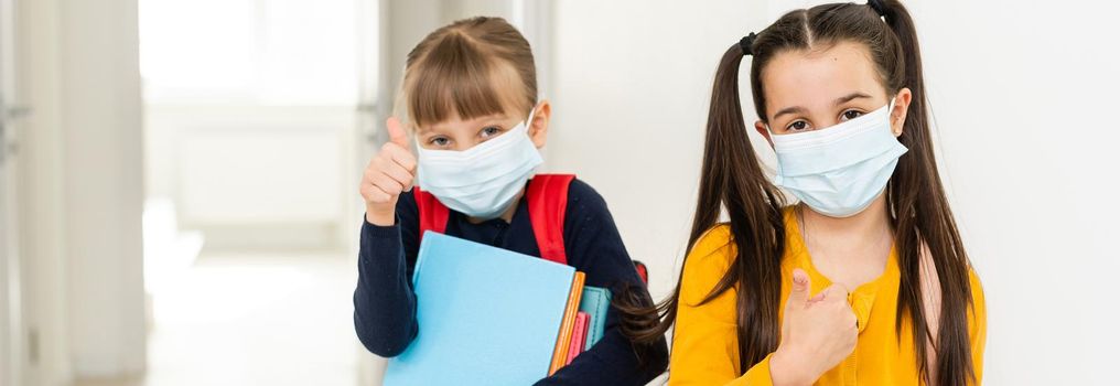 Cute little girls of primary hug each other with the mask on. School girls carry notebooks in hand and greet each other before entering school