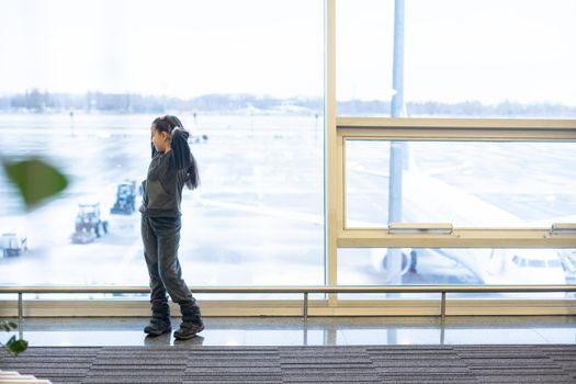 little baby girl waiting boarding to her flight in airport transit hall and looking at airplane parking near departure gate. Travelling by air with child.