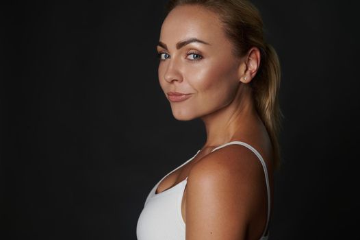 Close-up portrait of a beautiful slim body Caucasian woman, fit and muscular build young fitness girl in white top confidently looking at camera, isolated over black background with copy ad space