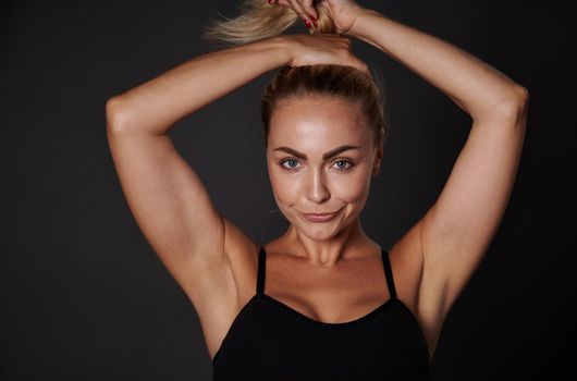 Sexy middle aged woman with toned body, beautiful healthy tanned skin, posing in black lingerie and tying a ponytail on her head, three quarter studio portrait over dark background with copy space