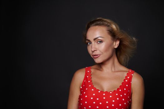 Close-up beauty portrait of an attractive gorgeous middle aged European woman in red top with white polka dots confidently looking at camera isolated over black background with copy ad space