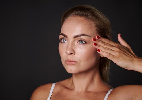 Close-up beauty portrait of a beautiful 30 years old blonde Caucasian woman with glowing fresh healthy skin holding her hand on her temple, isolated over black background with copy space