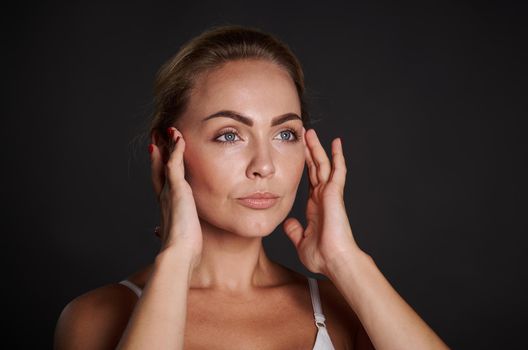 Close-up portrait of an European pretty woman with fresh glowing skin holding hands on her temples, doing massage movements on her face. Anti-aging concept, smoothing, rejuvenating beauty treatment