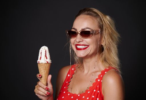 Cheerful middle aged blonde woman wearing protective sunglasses and red swimsuit, holding a cornet ice-cream and smiles toothy smile looking at camera, isolated over black background with copy space