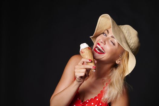 Attractive European blonde woman in summer hat holding a delicious ice-cream in her hands and smiles with beautiful toothy smile looking at camera, isolated over black background with copy ad space