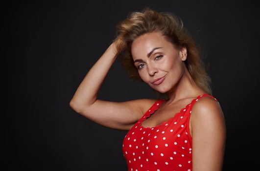 Attractive provocative sexy blonde woman in red swimsuit holding her hand on head and touching her flying healthy shiny hair, looking at camera isolated over black background with copy ad space