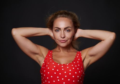 Attractive blonde middle aged Caucasian woman in red swimsuit with white polka dots confidently looking at camera posing holding hands behind her head against black background with copy ad space