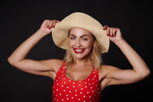 Attractive Caucasian blonde pretty woman in a straw summer hat and red swimsuit with white polka dots smiling with beautiful toothy smile standing against black background with copy ad space