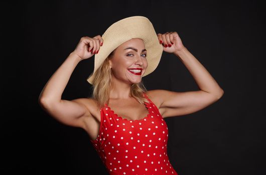 Attractive Caucasian blonde woman in a red swimsuit with white polka dots and a straw summer hat smiles a beautiful toothy smile, looking at the camera posing against black wall background. Copy space