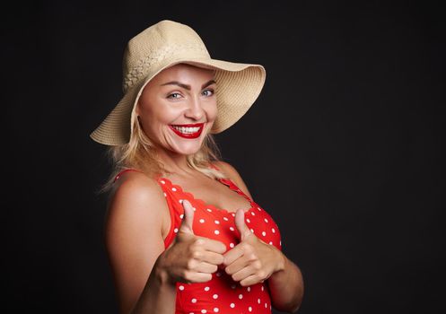 Delightful blonde Caucasian woman in summer straw hat and red swimsuit winking and smiling with beautiful toothy smile, showing thumbs up at camera, posing over black background with copy ad space