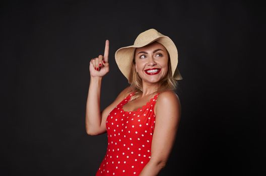 Happy cheerful stunning blonde Caucasian woman in red outfit with white polka dots and straw summer hat smiles beautiful white toothy smile, pointing up finger at copy space on black background